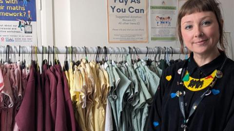 A woman is standing to the right of the drame. She is wearing a black shirt and has her hair tied up. Next to her is a rail with school uniforms in different colours hanging from it.