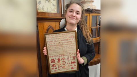 Auctioneer holding the sampler. It sits in a brown portrait frame, and is around 2 feet long and a foot wide. The stitching is red and shows various fonts of stitched letters from A to Z as well as flower pattern motifs and images, including a house and a steam engine.