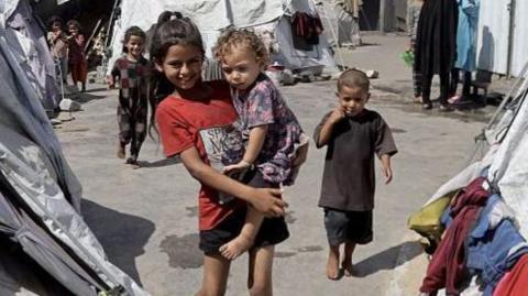 A smiling older child carries a younger girl in her arms in a displaced persons camp in Khan Younis, Gaza. The pair are surrounded by dirty grey and white tents with items of clothing drying on the outside. there are other children and  women in background.
