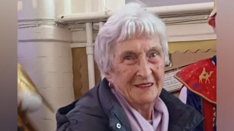 Rose Allan is looking just past the camera and smiling slightly. She has short white hair and is wearing a black coat and a lilac scarf.