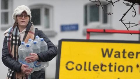 A woman in a hat carrying several water bottles beside a yellow water collection sign