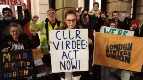 Shauni-Leigh Tyson standing outside a council building surrounded by a crowd of protesters, holding a placard reading 'Cllr Virdee: Act Now!'. The other protesters are also holding placards and flags with messages like 'Mould affected my health', and they are all raising their fists.