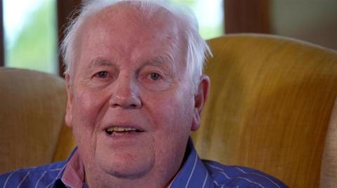 A picture of Sir David Kirch. He is smiling at the camera wearing a striped blue shirt. He sits on a yellow sofa.