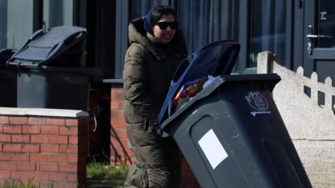 A woman in a long green puffa coat, sunglasses and pink trainers wheeling a grey bin along the pavement.