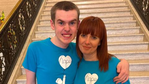 A boy with dark hair has his arm around a woman with dark hair, they are both wearing blue and white tops.