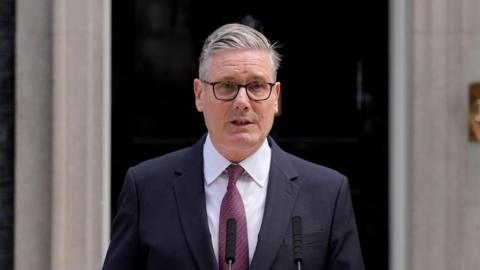 Sir Keir Starmer wearing a dark suit and tie standing outside 10 Downing Street. He is giving a speech from a lectern.