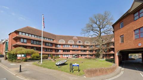 The outside of Surrey Heath Borough Council in Knoll Road, Camberley. It is a multi-storey brick building around the outside of an area of grass.