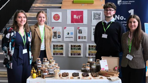 The three women and one man stand by a table with their snacks and product information on. A board is visible behind them with small posters one and they are stood by some stairs to the left.