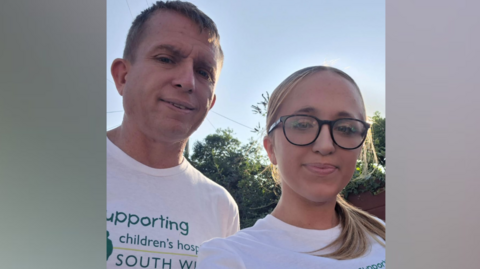 Freya Bird is pictured with her dad. Both are looking directly at the camera and they are wearing white charity T-shirts. The logo on their T-shirts says 'I'm supporting Children's Hospice South West'.