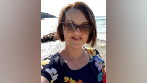 Chloe is smiling in a selfie she is taking at a beach. She is wearing sun glasses and a floral print top.