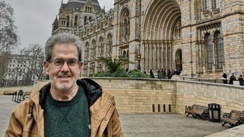 Donald Mitchell, who has grey hair and glasses stood outside a grand building wearing a green jumper and brown jacket.