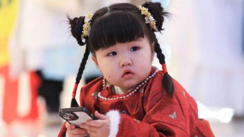 This photo taken on 19 November 2023 shows a child wearing a red Chinese traditional dress, known as Hanfu, looking at a smartphone during a Hanfu parade in Shenyang in China's northeastern Liaoning province. 