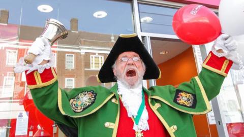 Jonathan Jones, 75, wearing his green town crier coat with a red shirt and black hat. He is holding a bell in his right hand and balloons in his left hand while shouting.