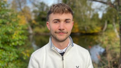 Michael Gamble stands outdoors in front of trees, bushes and a pond. He is wearing a light-coloured zip-up sweater with a small embroidered logo on the chest. Yellow leaves among the greenery suggest an autumn setting in a park.