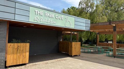 The exterior of The Park Cafe in Exhibition Park, which had a pastel green sign with the outline of a swan on it. The shutters are closed over the entrance. There are planters either side of the entrance and benches underneath an outdoor canopy made from wood.