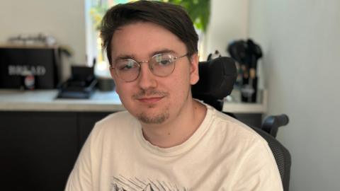 Jack is wearing glasses and a cream T-shirt. He is sitting in his wheelchair in the kitchen