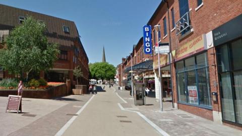 Alcester Street in Redditch with white lines painted on the side of the road, shops on the right and an open area with a walled garden and large tree to the left. There is a church spire in the distance behind a large bushy green tree.