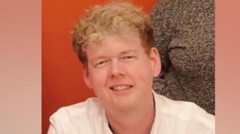 A man with blonde hair. He is looking at the camera and smiling. Behind him is an orange background. 