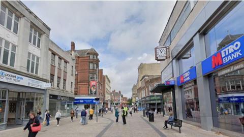 A view of a busy shopping street, pedestrians are seen walking throughout, there are rows of shops on either side and to the right is a building with a sign that says Metro Bank