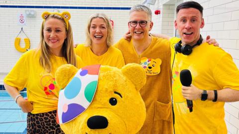 The four BBC News NI radio presenters wearing a range of Pudsey themed clothing. Cate Conway wears a yellow t-shirt, leopard print bottoms and a Pudsey ear headband.  Anne-Marie has short blonde hair and wears a yellow t-shirt, Mark Simpson wears a yellow fleece with a Pudsey badge. And finally on the right Connor Phillips wears a yellow t-shirt with earphones around his neck and a microphone in his hand.