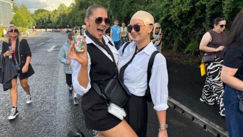 Two women. Both are wearing white shirts, black ties and sunglasses. The one on the right is wearing a bald cap and the one on the left is wearing a black waiscoat.
