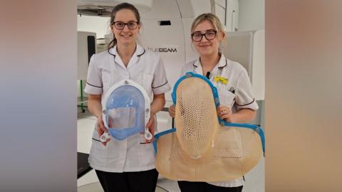 Two nurses are standing next to each other. On the left the woman has brown hair and glasses, and is wearing a white uniform. On the right the woman has blonde hair and glasses, with white uniform and a yellow badge. On the left the woman is holding a small mask that goes over someone's face - it has blue mesh that covers much of the patient's face but with a large hold for their nose and mouth. On the right, the mask is mesh and would cover the entirety of someone's head and shoulders