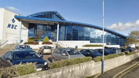 The exterior of the NSC, which is a large glass fronted building with a curved blue roof and a lettering that reads NSC a white wall next to the entrance. There are cars parked in front of the building.