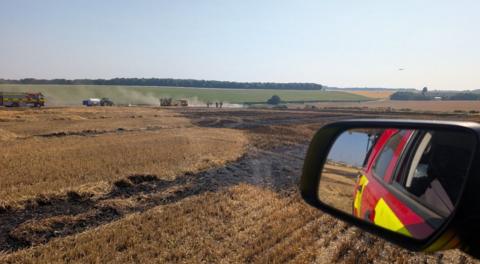 In the distance you can see small puffs of smoke drifting up from a farmer's field. The fire is all but you can also see a fire engine parked up on the edge of the field which is bone dry aside from the black patches on the ground where the fire has burned. In the foreground you're looking into the wing mirror of a fire service 4x4 vehicle, leaving you looking back at the side of the car itself and at more parched fields with more patches of black dotted around.