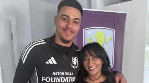 Morgan Rogers on the left has his left arm around Zena Weeks and they are both smiling at the camera. A large Aston Villa crest is behind them.