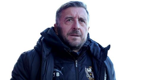 Port Vale manager Jon Brady walks along the touchline in his club tracktop and jacket