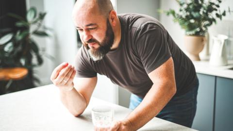 A man leans over a counter with a glass of water and a tablet