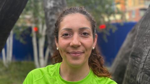 A woman - Renata Wais - smiling directly at the camera. She is standing in front of a wooden sculpture and is wearing a green running T-shirt.