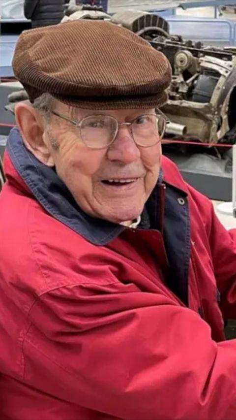 Elderly man wearing a brown cap and glasses and red jacket sat outdoors