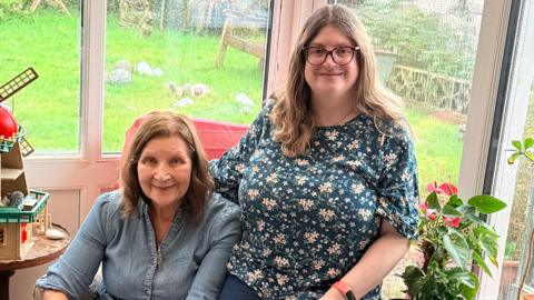 Sarah Edgeworth is perched on the edge of a chair. She has long brown hair, glasses and is wearing a floral top. She is next to her mum Sue, who is sitting on the chair and smiling. She has short brown hair and is wearing a blue top.
