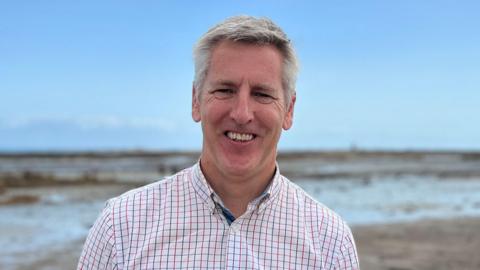Helier is in a white checkered shirt and dark trousers stands smiling on a beach with a rocky path leading toward the sea. The background features wide sandy flats, shallow water, and a partly cloudy sky.