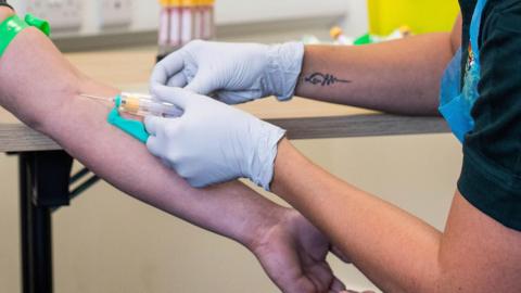 A nurse uses a needle to take blood from a patient's arm. The nurse is wearing white gloves, a green short-sleeved top and a blue disposable apron. The patient has a green tourniquet around their upper arm. They are sitting in a surgery with a wood-effect and black-metal table behind them. 