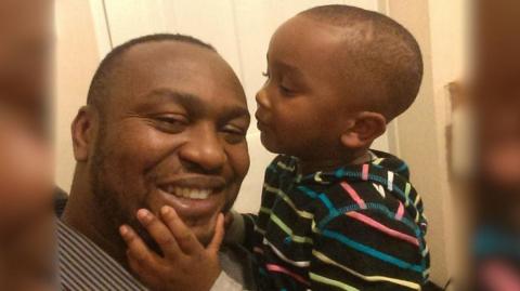 Chizi is shown being held by his father in their home. He is wearing a black top with colourful stripes. His father is wearing a striped shirt and is smiling. Chizi is about to kiss his dad on the cheek. 
