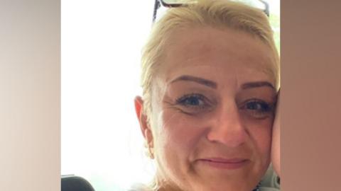 Beata Szauer, with an arm up to her face, with tied back blonde hair. She has a pair of glasses on her head. She is smiling at the camera. 
