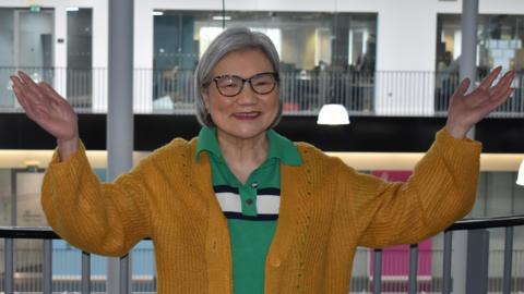 Kin Alderson wearing a mustard coloured cardigan over a green polo shirt and glasses. She is smiling and has her hands and arms aloft.