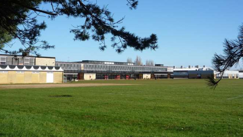 External view of Smithdon High School in Hunstanton. A two-storey building with lots of glass windows. In front of it is a green grass hockey pitch.