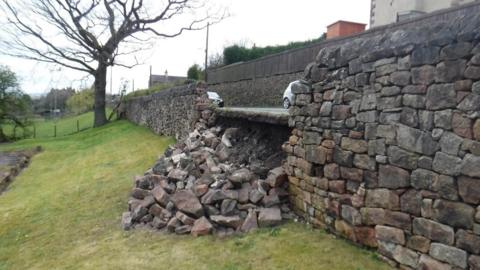 Part of a stone wall running along a road has collapsed on to the grass beneath it. Cars can be seen driving along the road in the background.