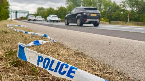 A piece of police tape is discarded on the grass verge in the foreground. Cars are travelling on the road beyond.
