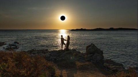 The ark shape of the Moon is in front of the sun, whose rays are shining out across the sea. The silhouettes of a couple are seen on rocks on the headland with the sea behind them and the sun's rays reflected in the water.