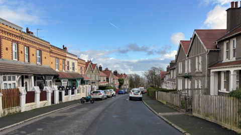 Street-view picture of Marathon Road. It's a blue sky with cars and motorbikes parked on the edge of the road. 