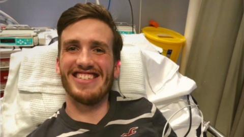 A young man with brown hair smiles as he lies in a hospital bed, with wires around him