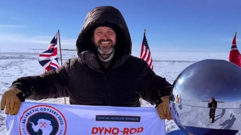Jordan Wylie, who has a dark beard with some white in it, stands smiling at the South Pole, next to a marker that looks like a mirrored ball. He has a black coat on with the hood up and is holding a sponsorship flag. In the background are  flags and a large flat expanse of ice under a blue sky