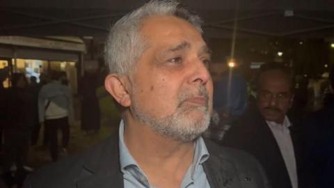 A middle aged man with grey hair and beard speaks to a reporter. It is night time and there are people in the background. 