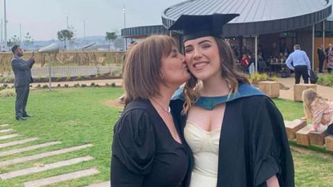 Sophie Morgan (right) pictured with her mother Erica in her cap and gown for her graduation. She smiles at the camera while her mother kisses her cheek. They are both stood outside. 