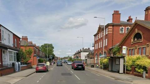 Google streetview of cars driving along Gorton Road past a bus shelter with houses and a civic centre on either side.