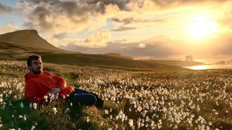 Man with beard and wearing red hoodie, dark trousers and yellow wellies, lying amid flowers in a field with hands clasped, and island coastline in background and sea, with sun glowing brightly in the sky.